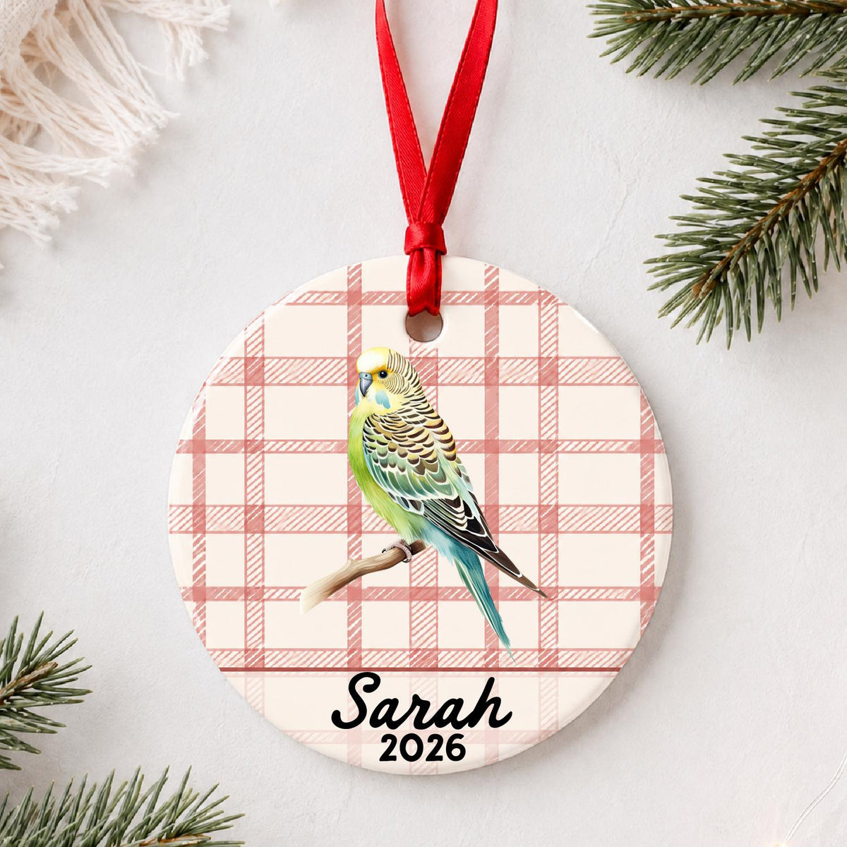 Personalized budgerigar ceramic ornament with name and year on red plaid styled with pine branches on a soft white background