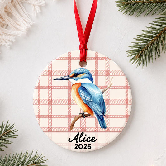 Personalized kingfisher ceramic ornament with name and year on red plaid styled with pine branches on a soft white background