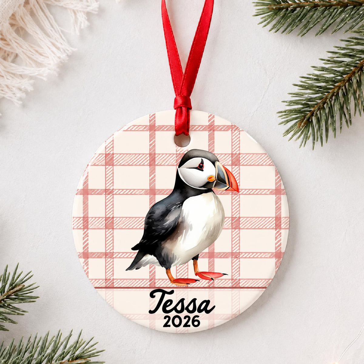 Personalized puffin ceramic ornament with name and year on red plaid styled with pine branches on a soft white background