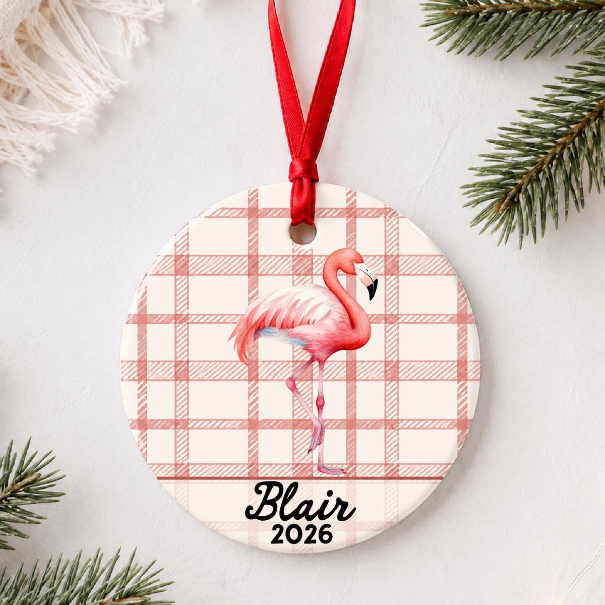 Personalized flamingo ceramic ornament with name and year on red plaid styled with pine branches on a soft white background