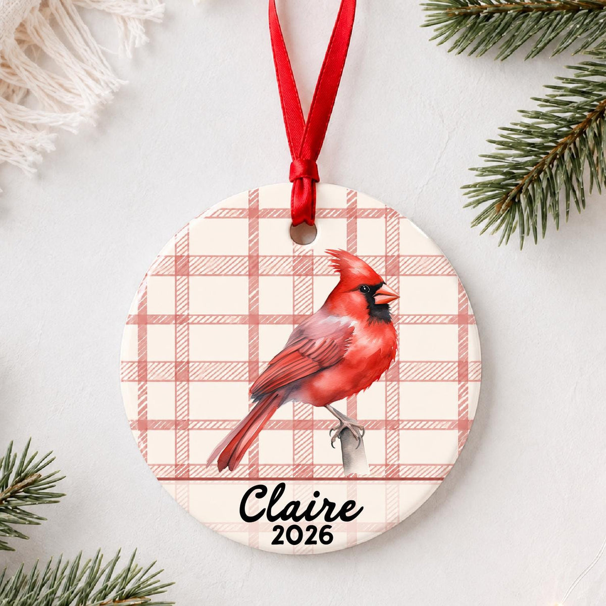 Personalized northern cardinal ceramic ornament with name and year on red plaid styled with pine branches on a soft white background