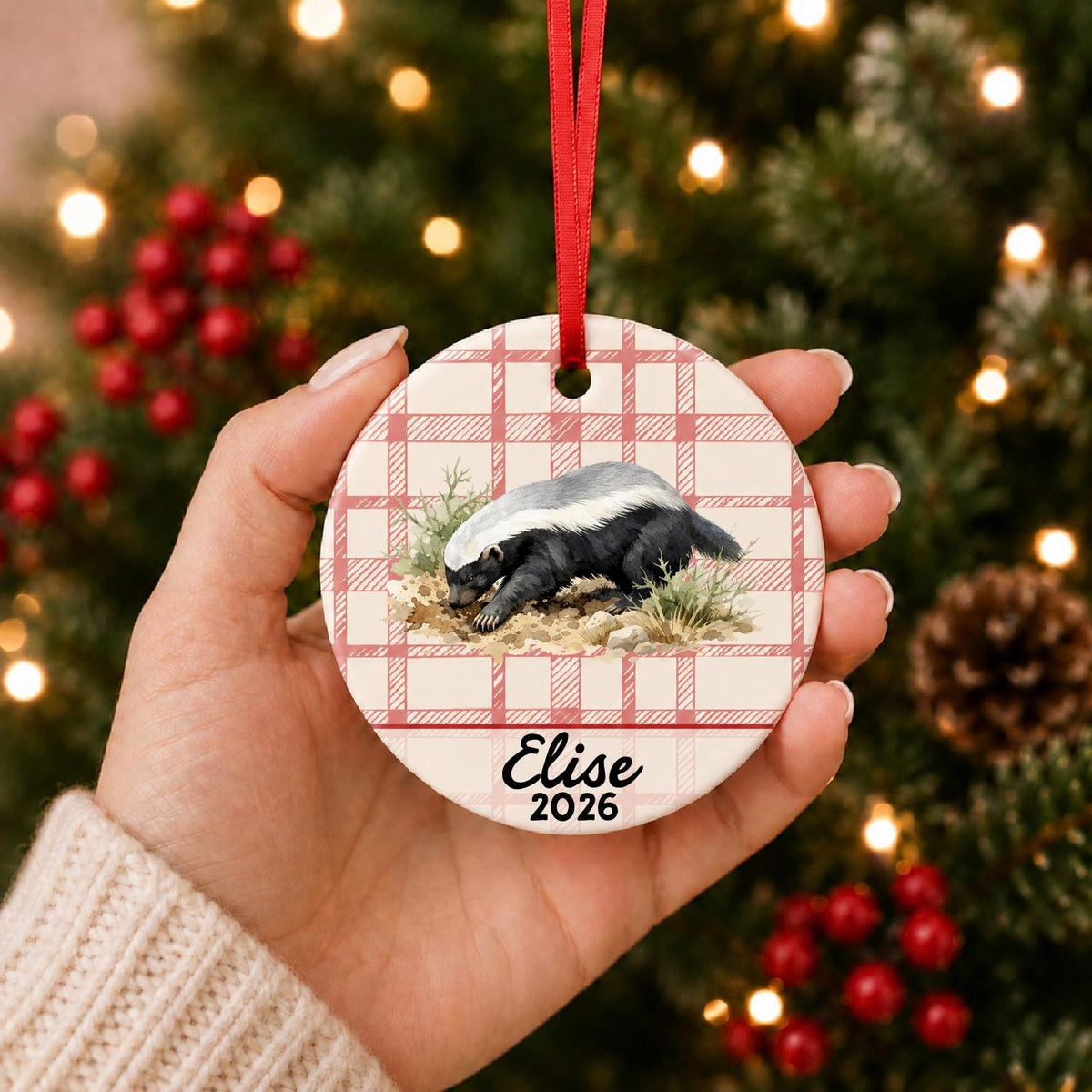Hand holding the personalized honey badger ceramic ornament with name and year on red plaid in front of a Christmas tree
