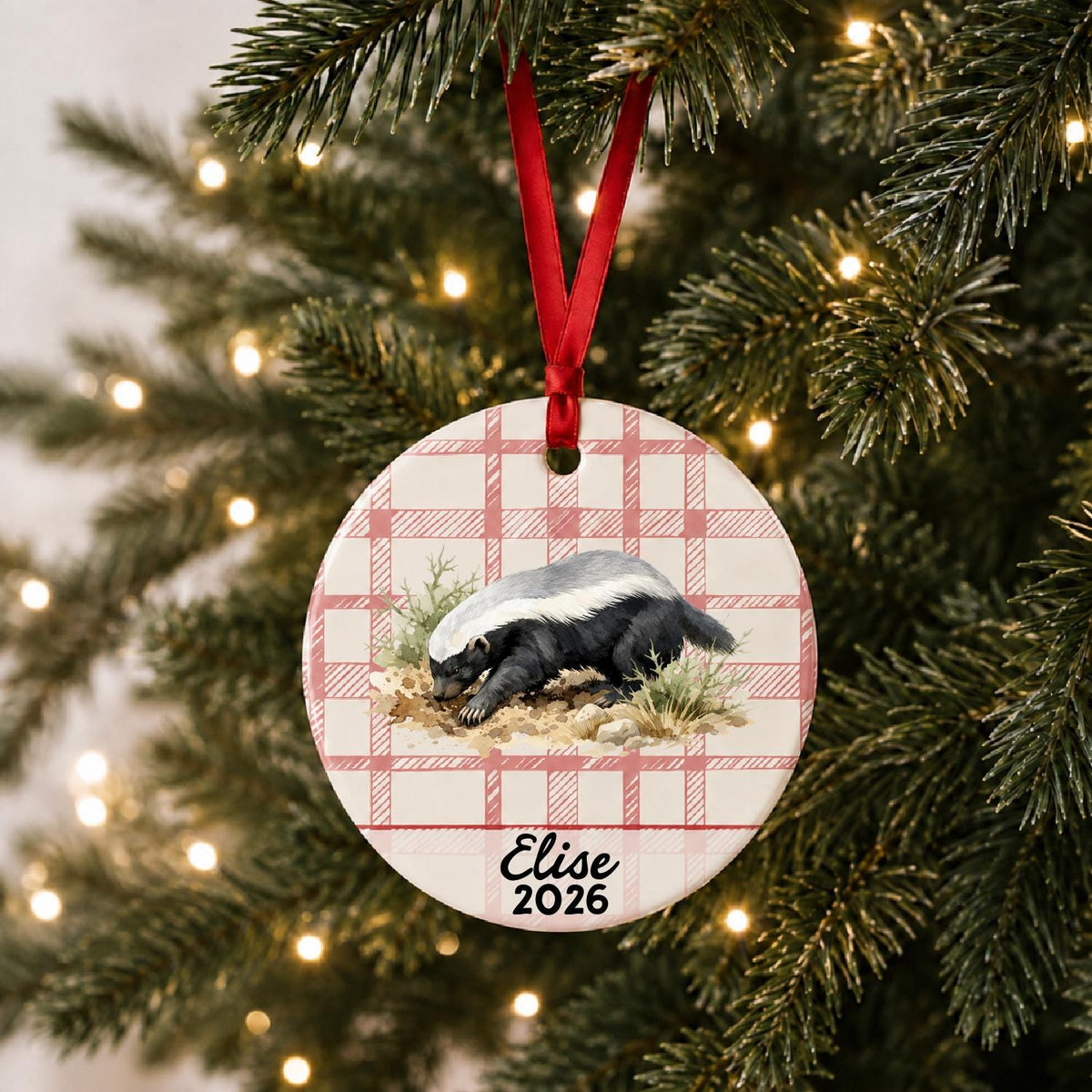 Personalized honey badger ceramic ornament with name and year on red plaid hanging on a lit Christmas tree