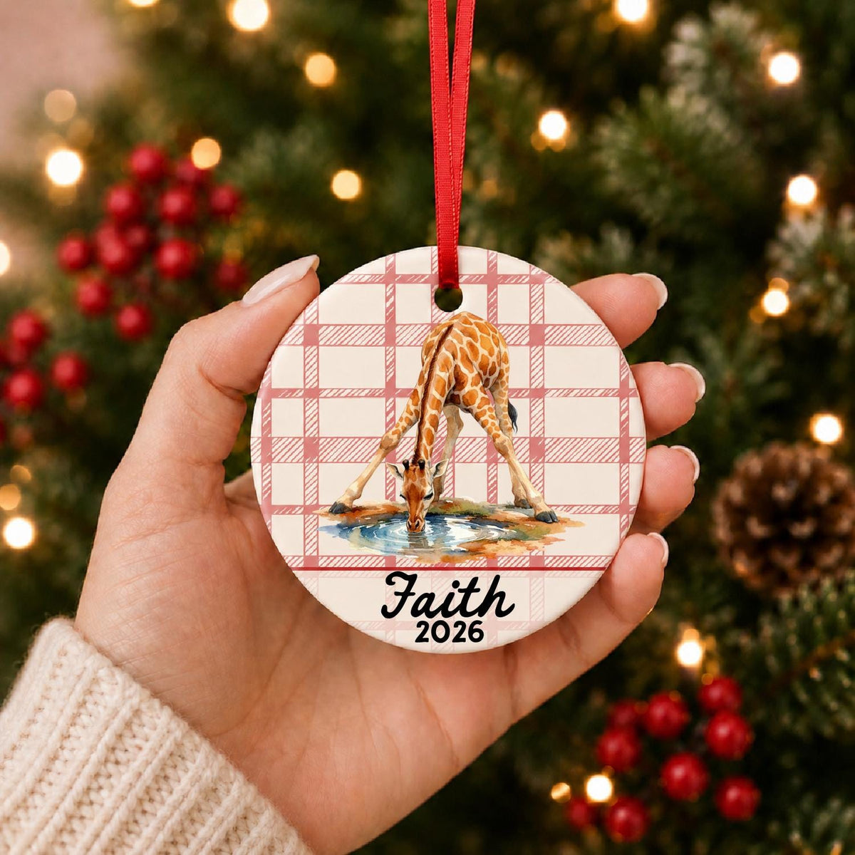 Hand holding the personalized giraffe ceramic ornament with name and year on red plaid in front of a Christmas tree