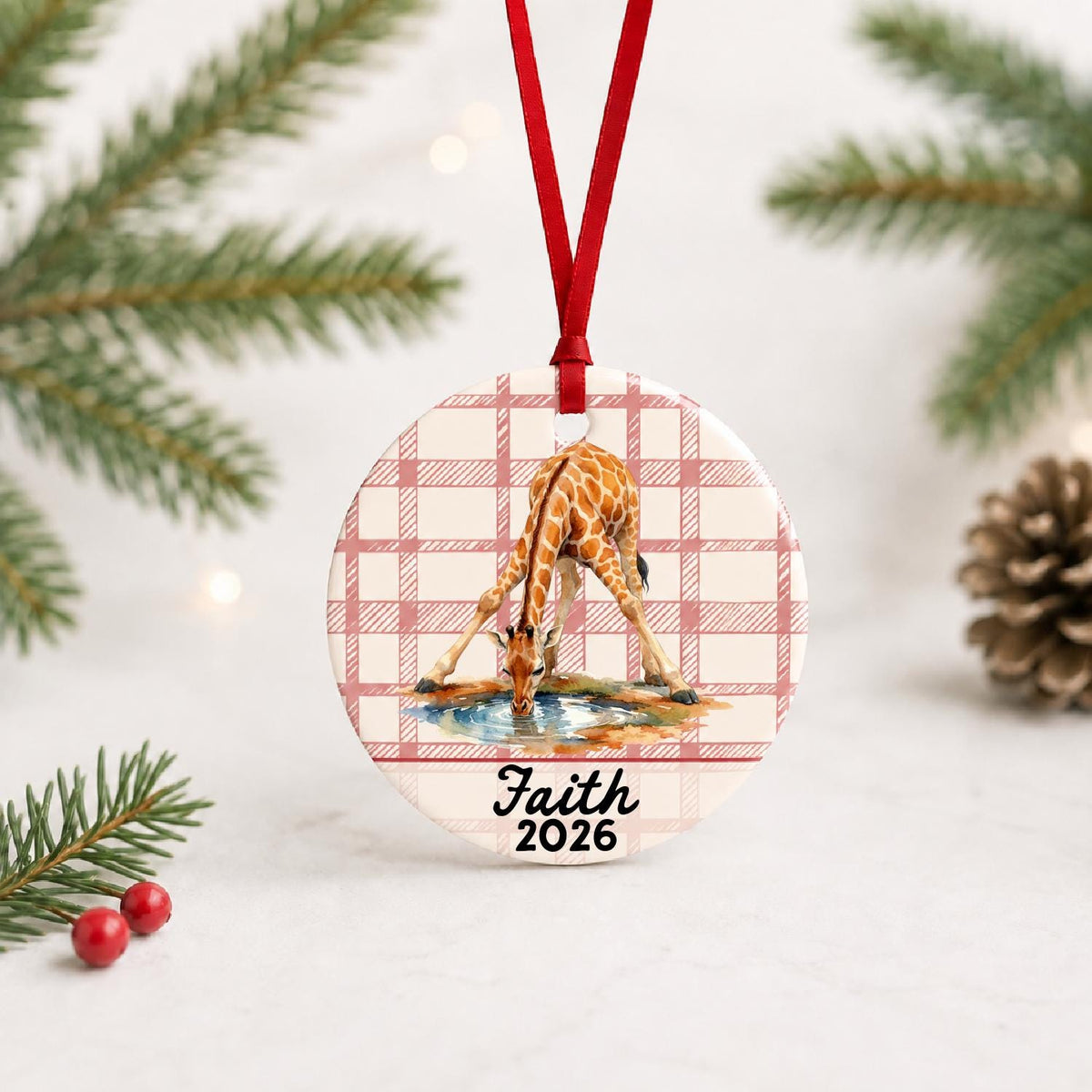 Personalized giraffe ceramic ornament with name and year on red plaid with red ribbon on a white surface with pine branches and pinecones
