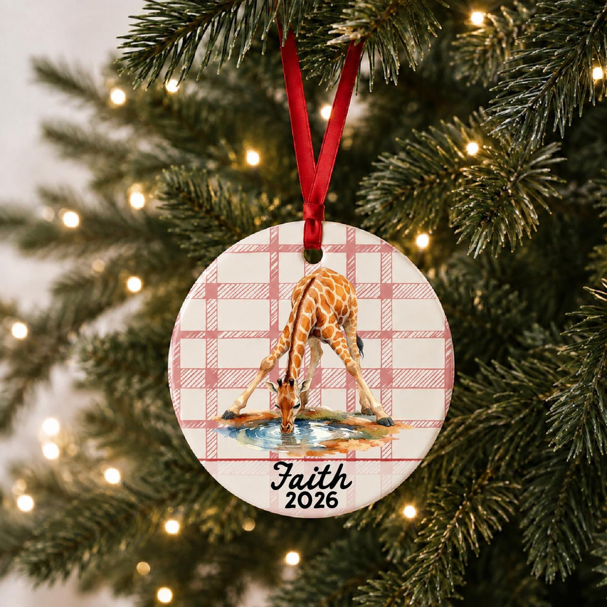 Personalized giraffe ceramic ornament with name and year on red plaid hanging on a lit Christmas tree
