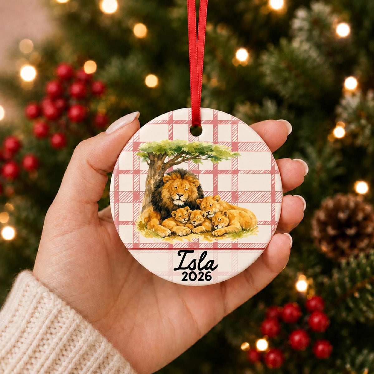 Hand holding the personalized lion pride ceramic ornament with name and year on red plaid in front of a Christmas tree