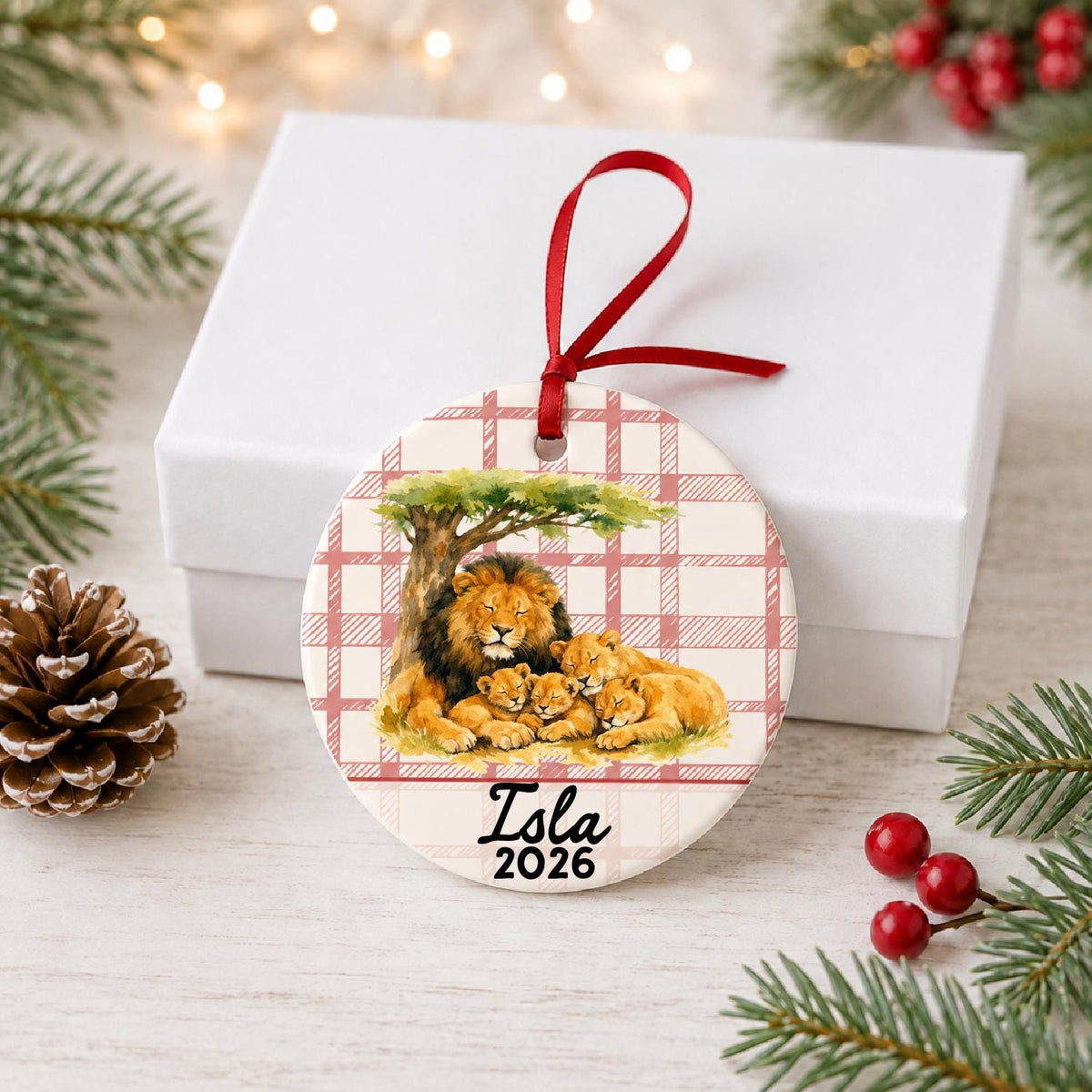 Personalized lion pride ceramic ornament with name and year on red plaid in front of a white gift box with pinecones and red berries