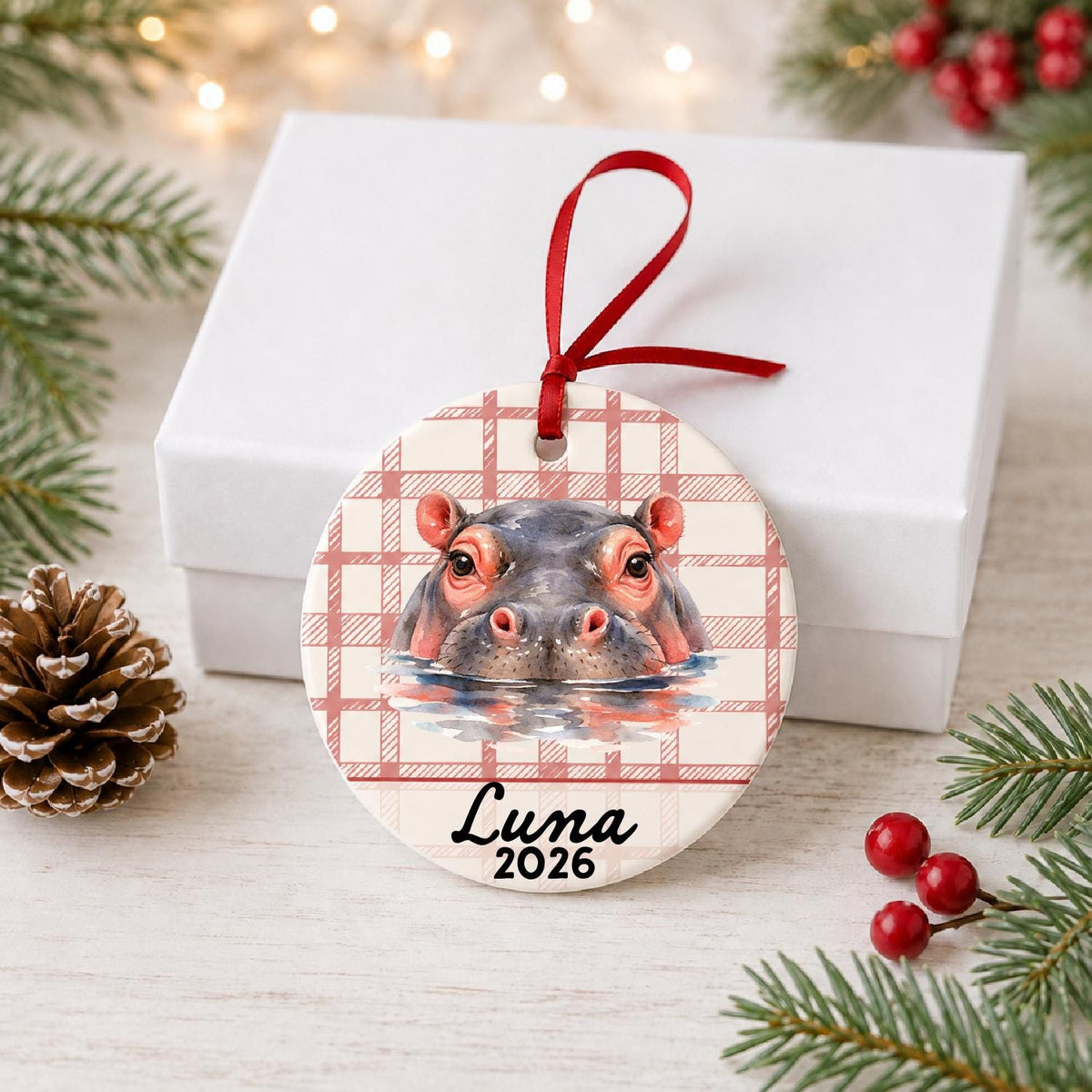 Personalized hippopotamus ceramic ornament with name and year on red plaid in front of a white gift box with pinecones and red berries