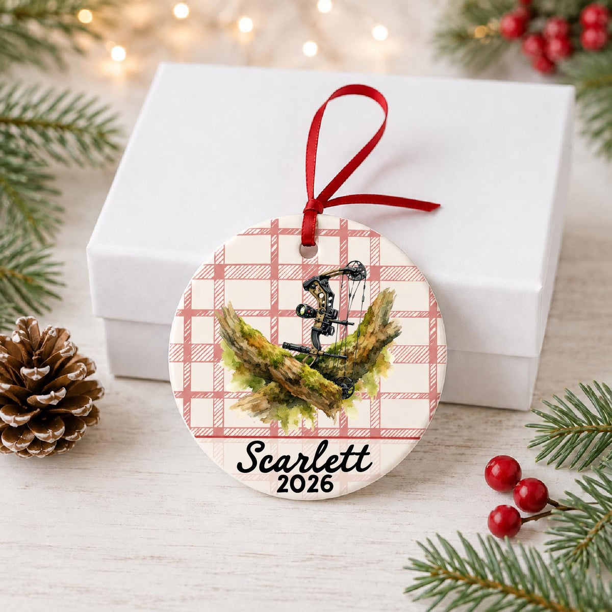 Personalized compound bow ceramic ornament with name and year on red plaid in front of a white gift box with pinecones and red berries