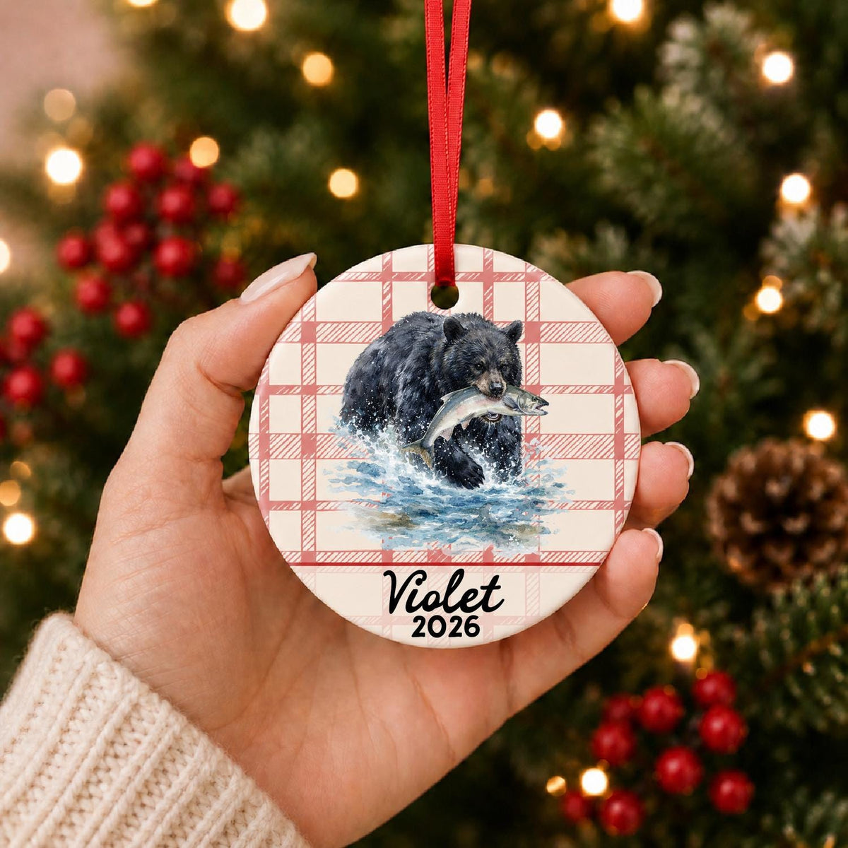 Hand holding the personalized American black bear ceramic ornament with name and year on red plaid in front of a Christmas tree