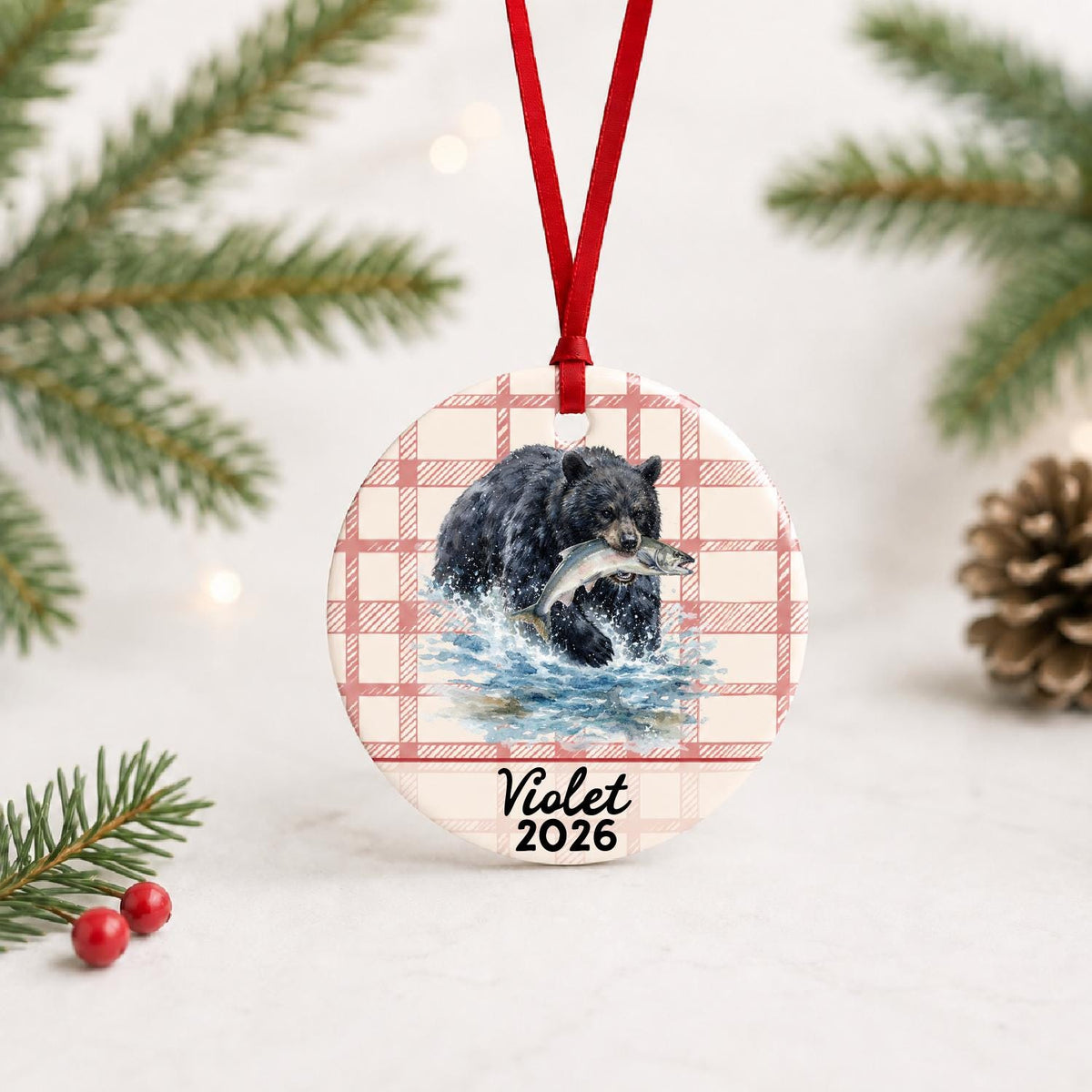 Personalized American black bear ceramic ornament with name and year on red plaid with red ribbon on a white surface with pine branches and pinecones