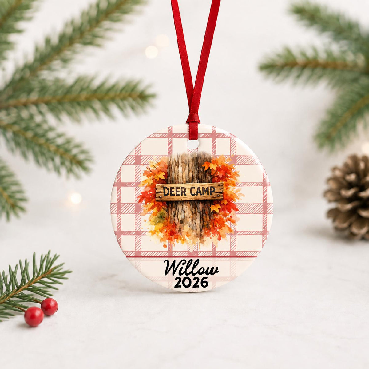 Personalized deer camp ceramic ornament with name and year on red plaid with red ribbon on a white surface with pine branches and pinecones