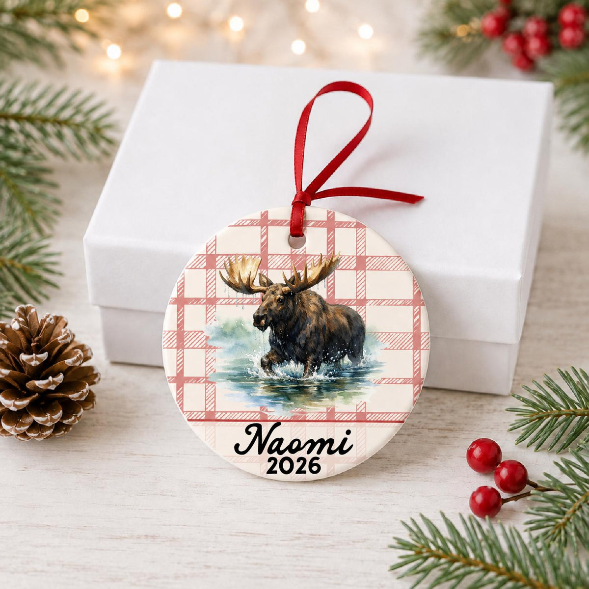 Personalized bull moose ceramic ornament with name and year on red plaid in front of a white gift box with pinecones and red berries