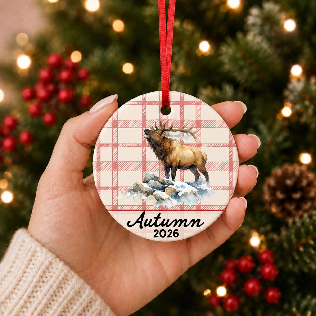 Hand holding the personalized bull elk ceramic ornament with name and year on red plaid in front of a Christmas tree