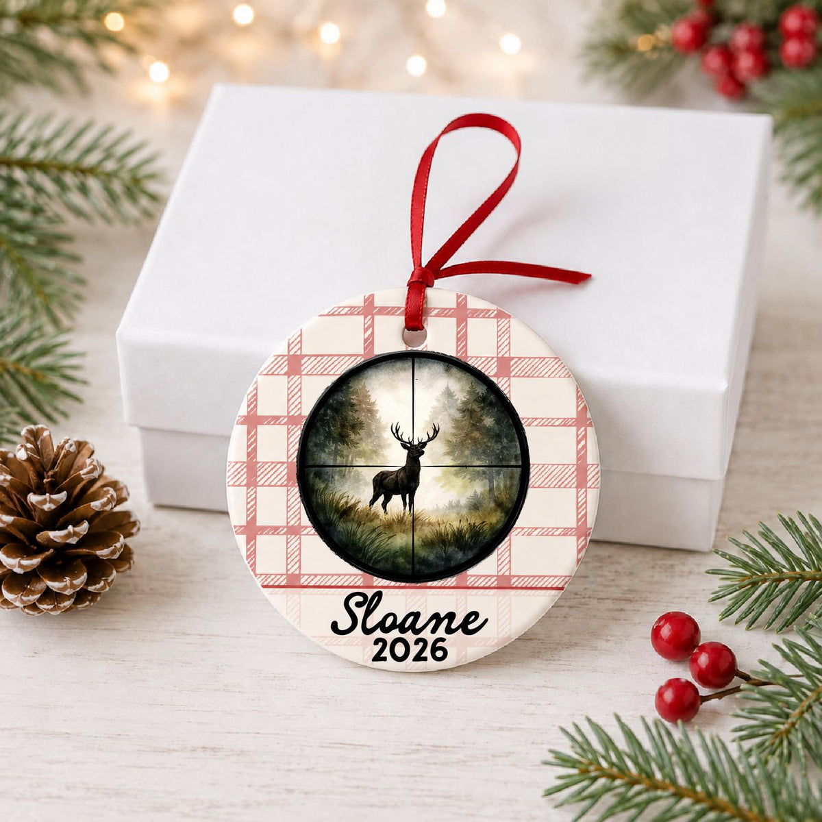 Personalized deer hunting ceramic ornament with name and year on red plaid in front of a white gift box with pinecones and red berries