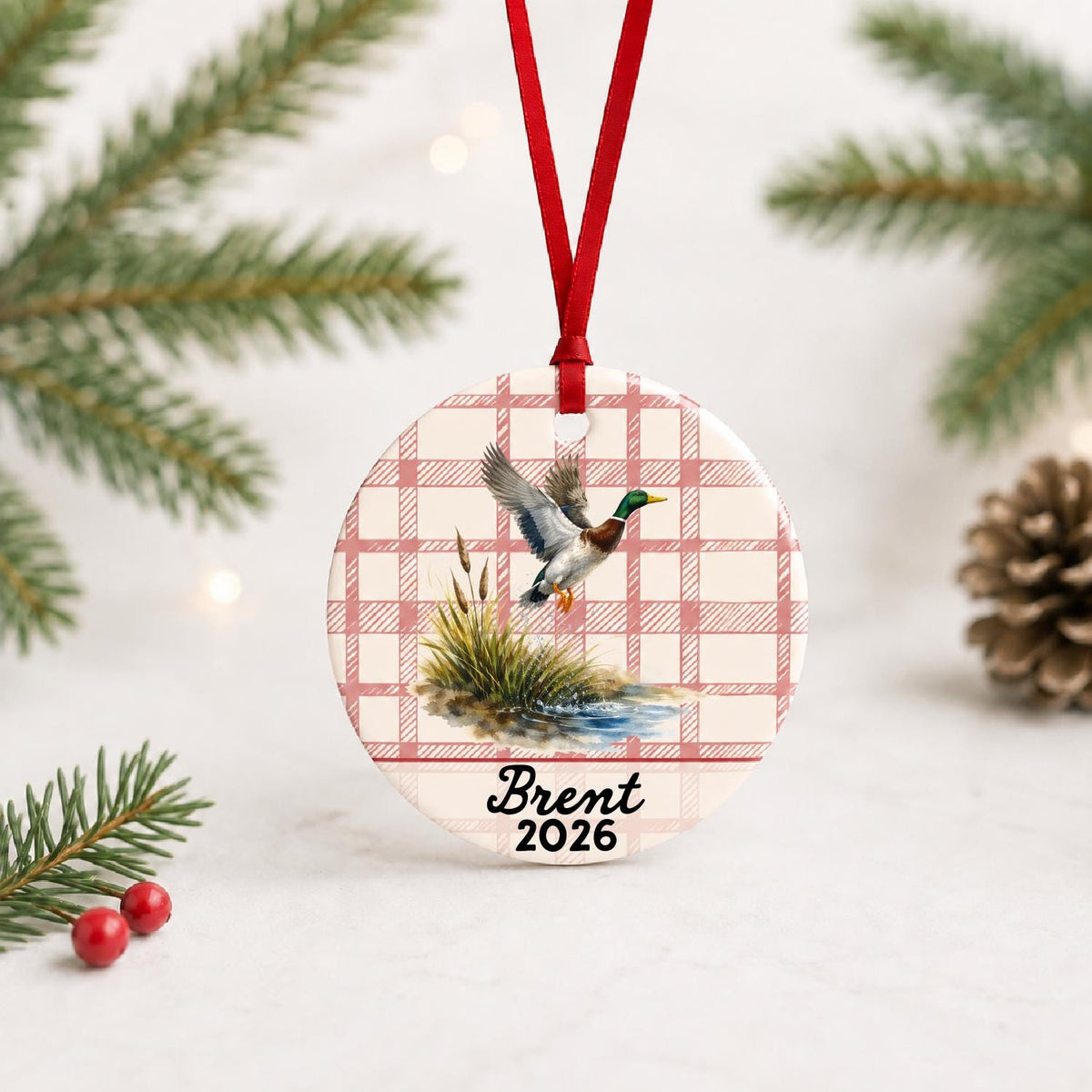 Personalized mallard duck ceramic ornament with name and year on red plaid with red ribbon on a white surface with pine branches and pinecones