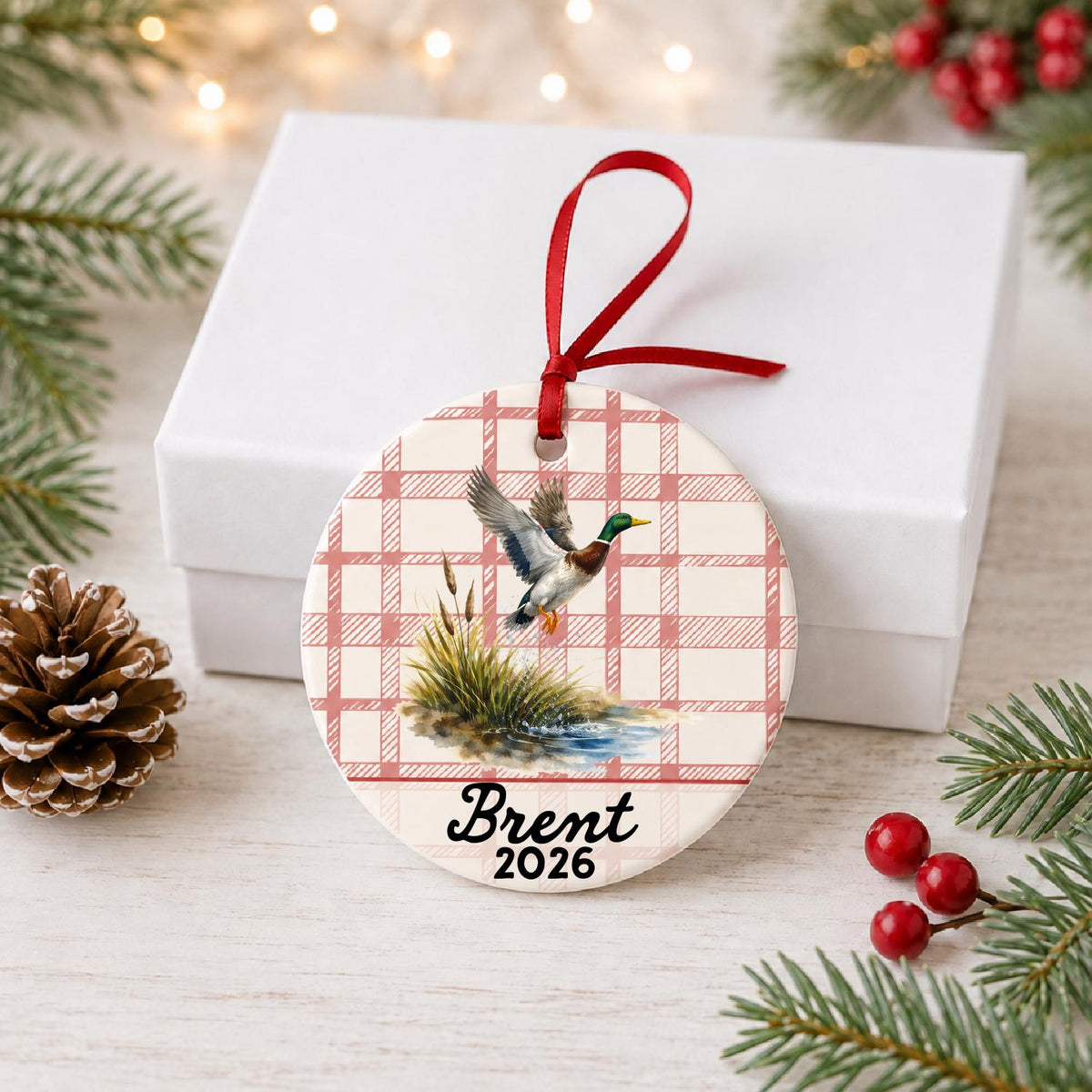 Personalized mallard duck ceramic ornament with name and year on red plaid in front of a white gift box with pinecones and red berries