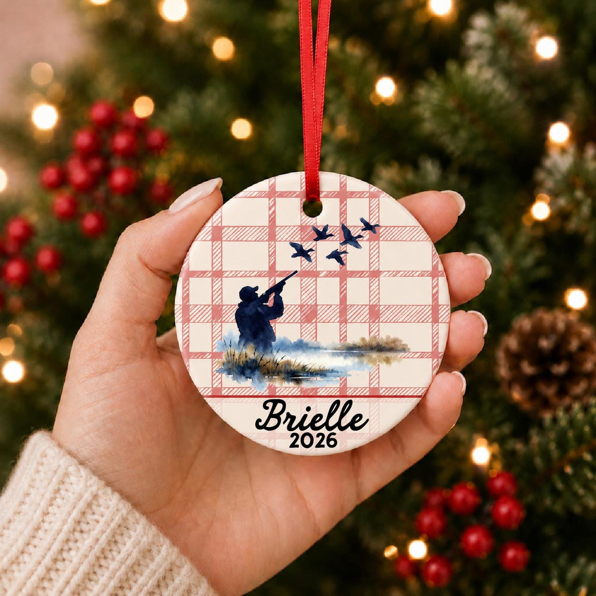 Hand holding the personalized duck hunting ceramic ornament with name and year on red plaid in front of a Christmas tree