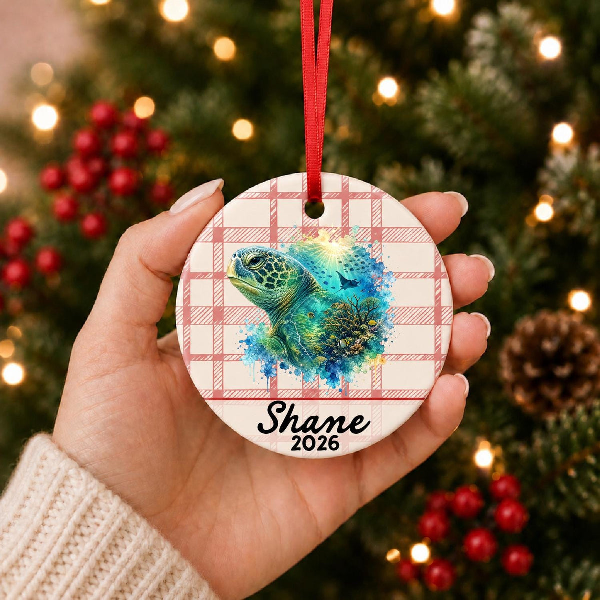 Hand holding the personalized green sea turtle ceramic ornament with name and year on red plaid in front of a Christmas tree
