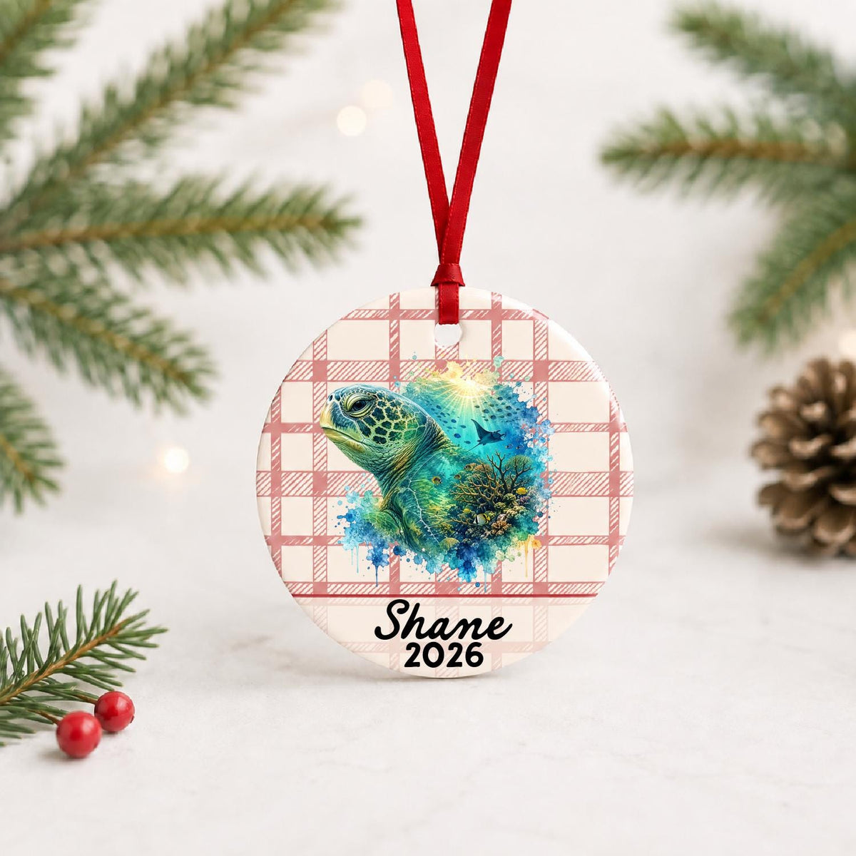 Personalized green sea turtle ceramic ornament with name and year on red plaid with red ribbon on a white surface with pine branches and pinecones