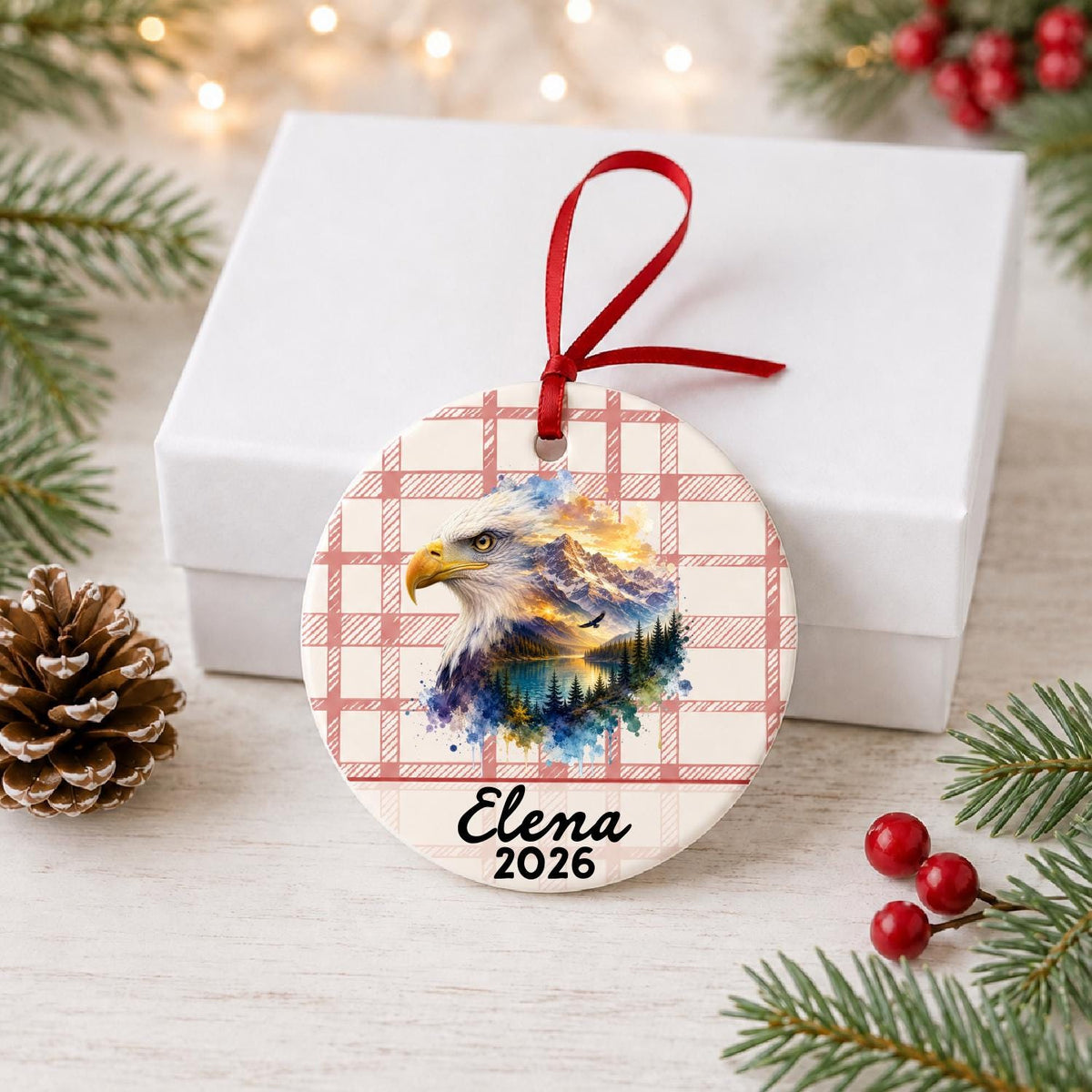 Personalized bald eagle ceramic ornament with name and year on red plaid in front of a white gift box with pinecones and red berries