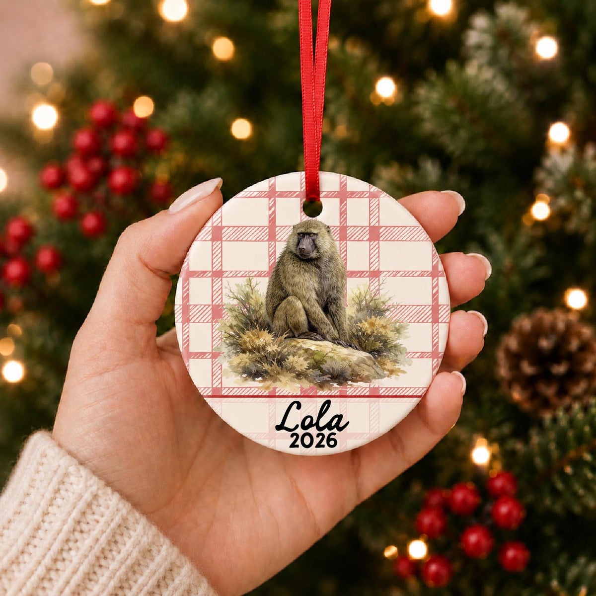 Hand holding the personalized baboon ceramic ornament with name and year on red plaid in front of a Christmas tree