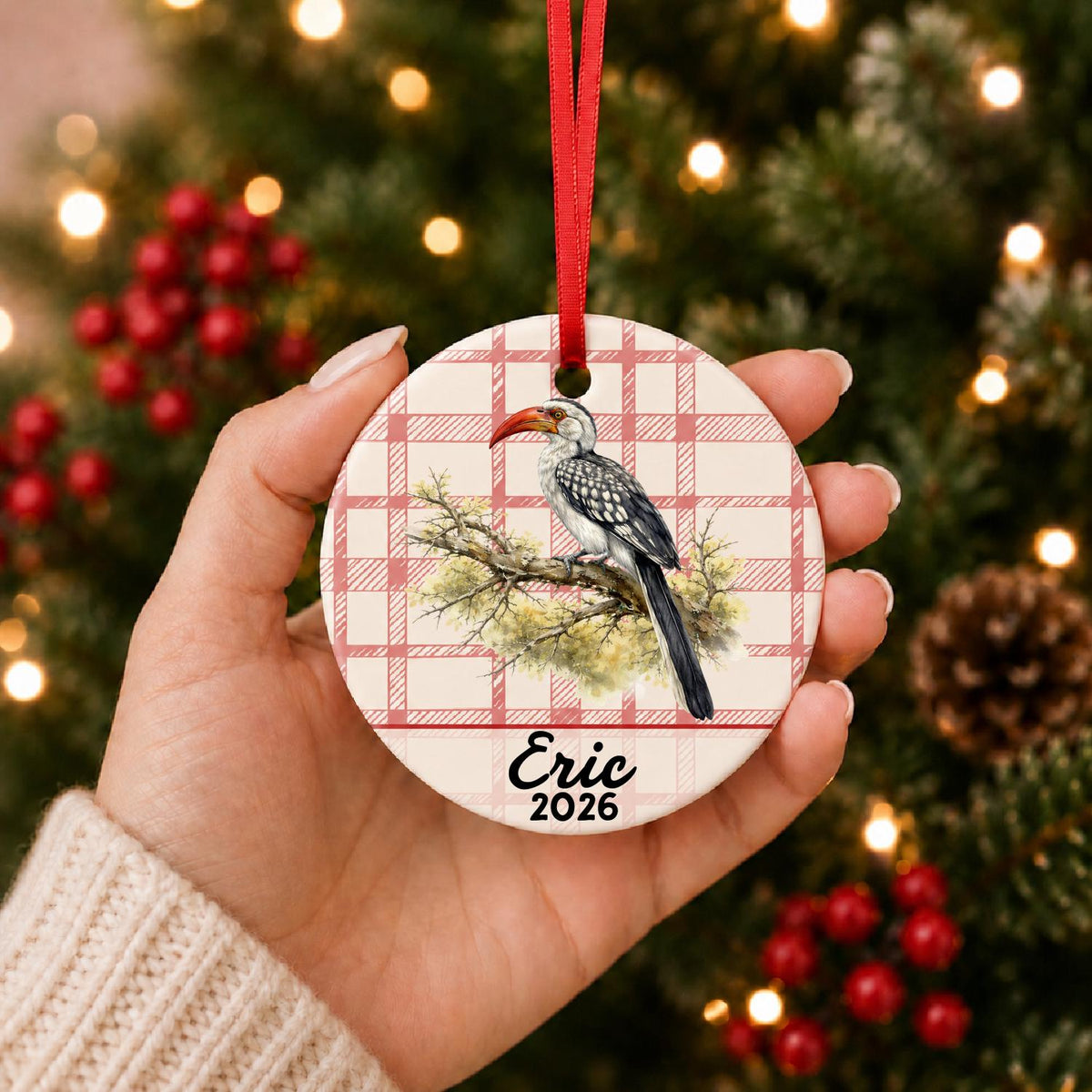 Hand holding the personalized hornbill ceramic ornament with name and year on red plaid in front of a Christmas tree