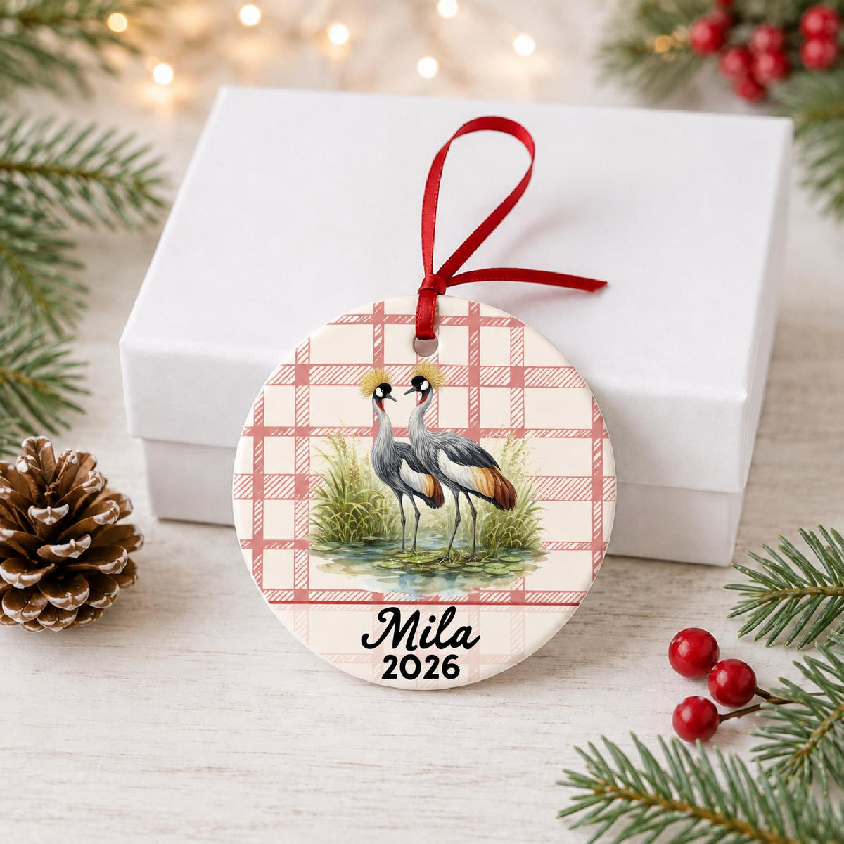 Personalized grey crowned crane ceramic ornament with name and year on red plaid in front of a white gift box with pinecones and red berries