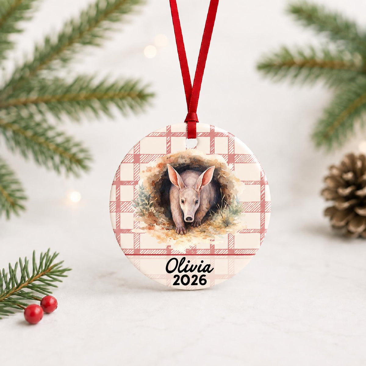 Personalized aardvark ceramic ornament with name and year on red plaid with red ribbon on a white surface with pine branches and pinecones