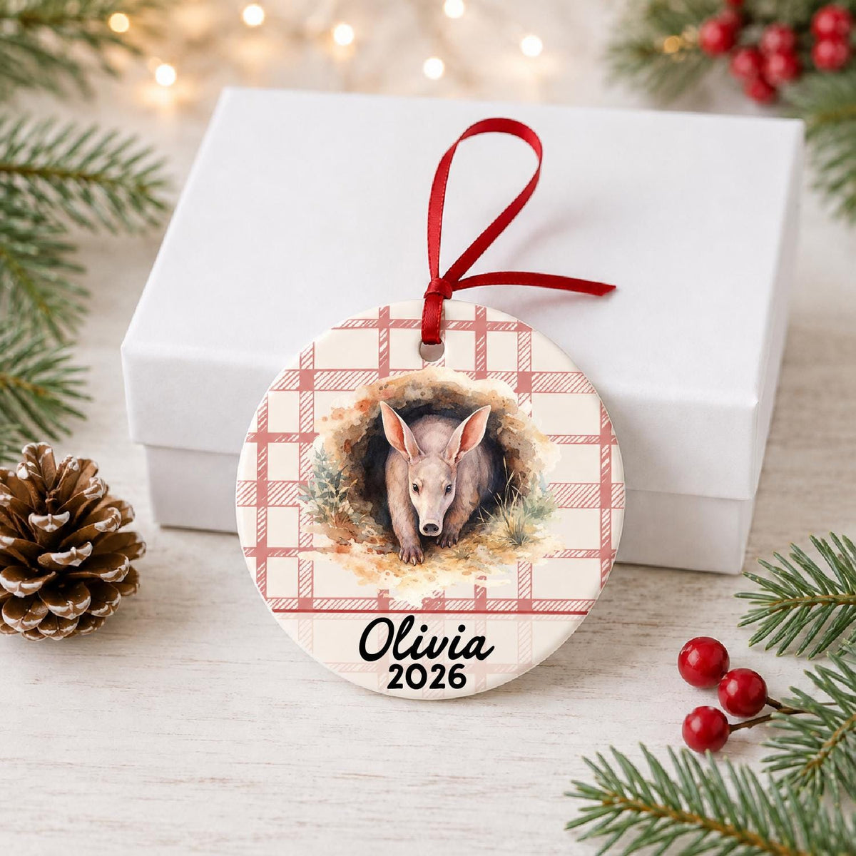 Personalized aardvark ceramic ornament with name and year on red plaid in front of a white gift box with pinecones and red berries