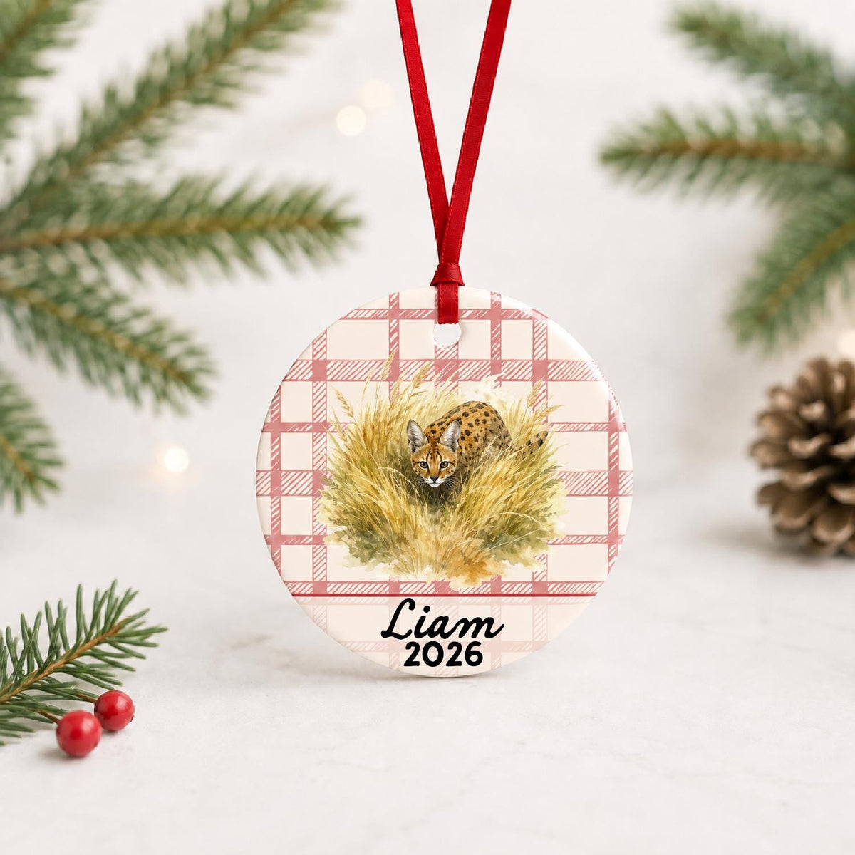 Personalized serval ceramic ornament with name and year on red plaid with red ribbon on a white surface with pine branches and pinecones