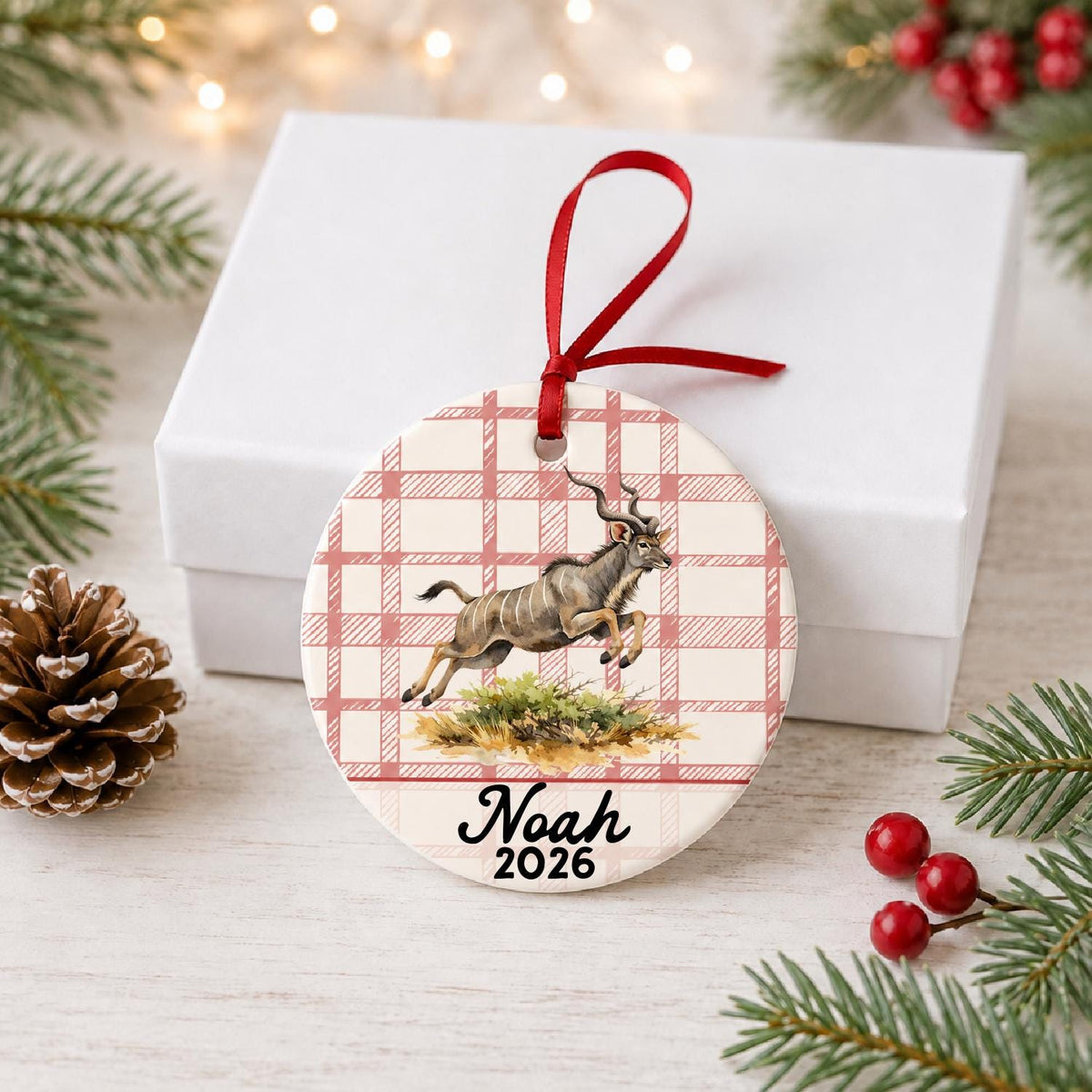 Personalized kudu ceramic ornament with name and year on red plaid in front of a white gift box with pinecones and red berries