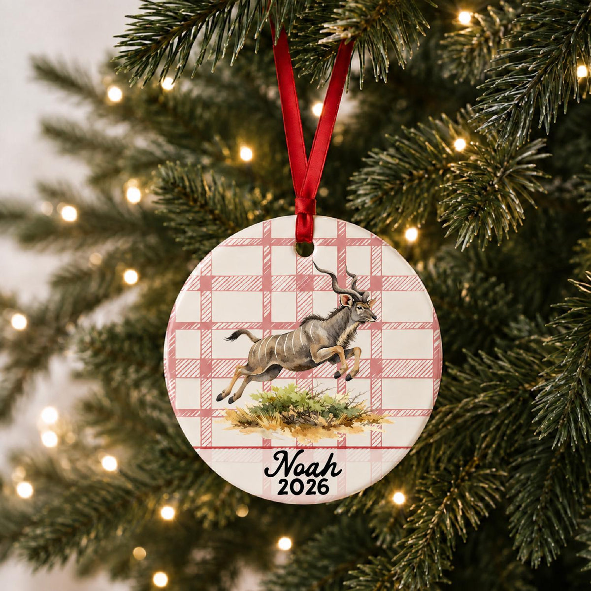Personalized kudu ceramic ornament with name and year on red plaid hanging on a lit Christmas tree