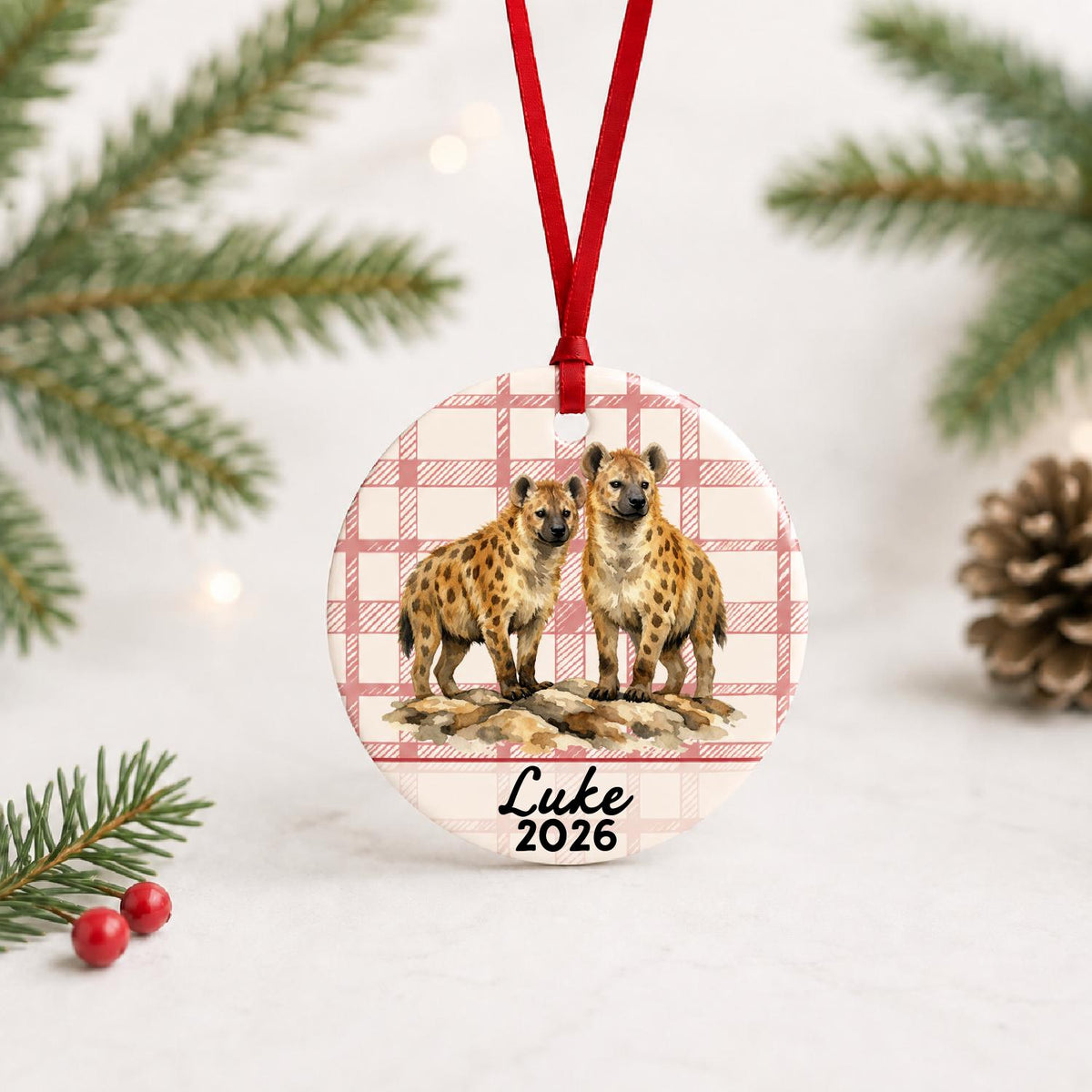 Personalized spotted hyena ceramic ornament with name and year on red plaid with red ribbon on a white surface with pine branches and pinecones