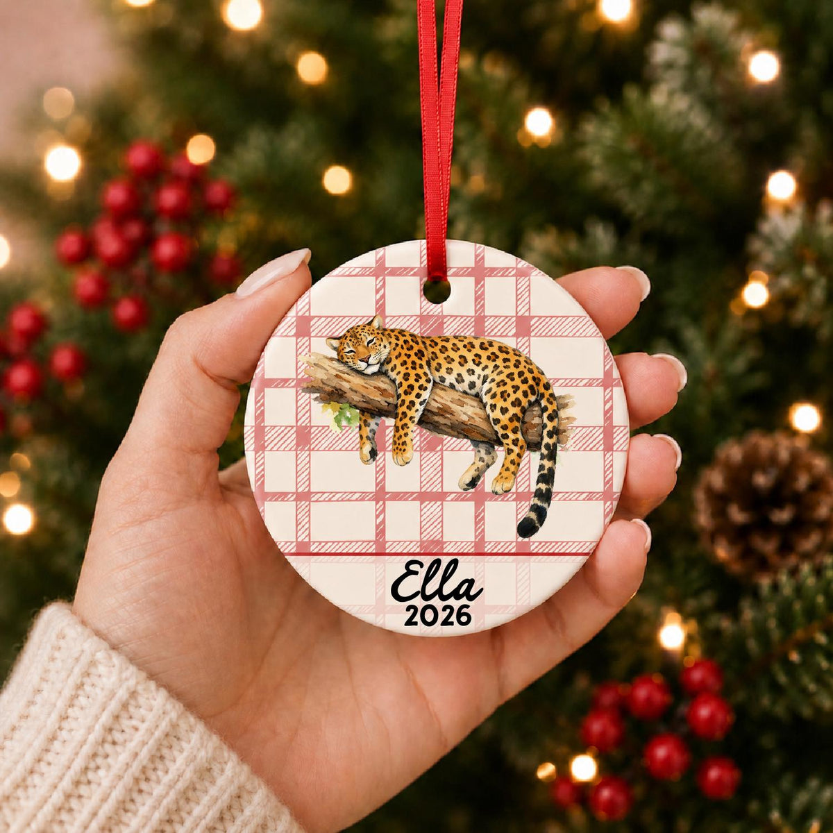 Hand holding the personalized leopard ceramic ornament with name and year on red plaid in front of a Christmas tree