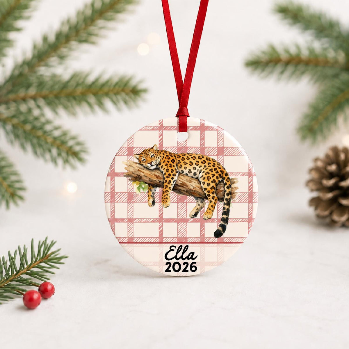 Personalized leopard ceramic ornament with name and year on red plaid with red ribbon on a white surface with pine branches and pinecones