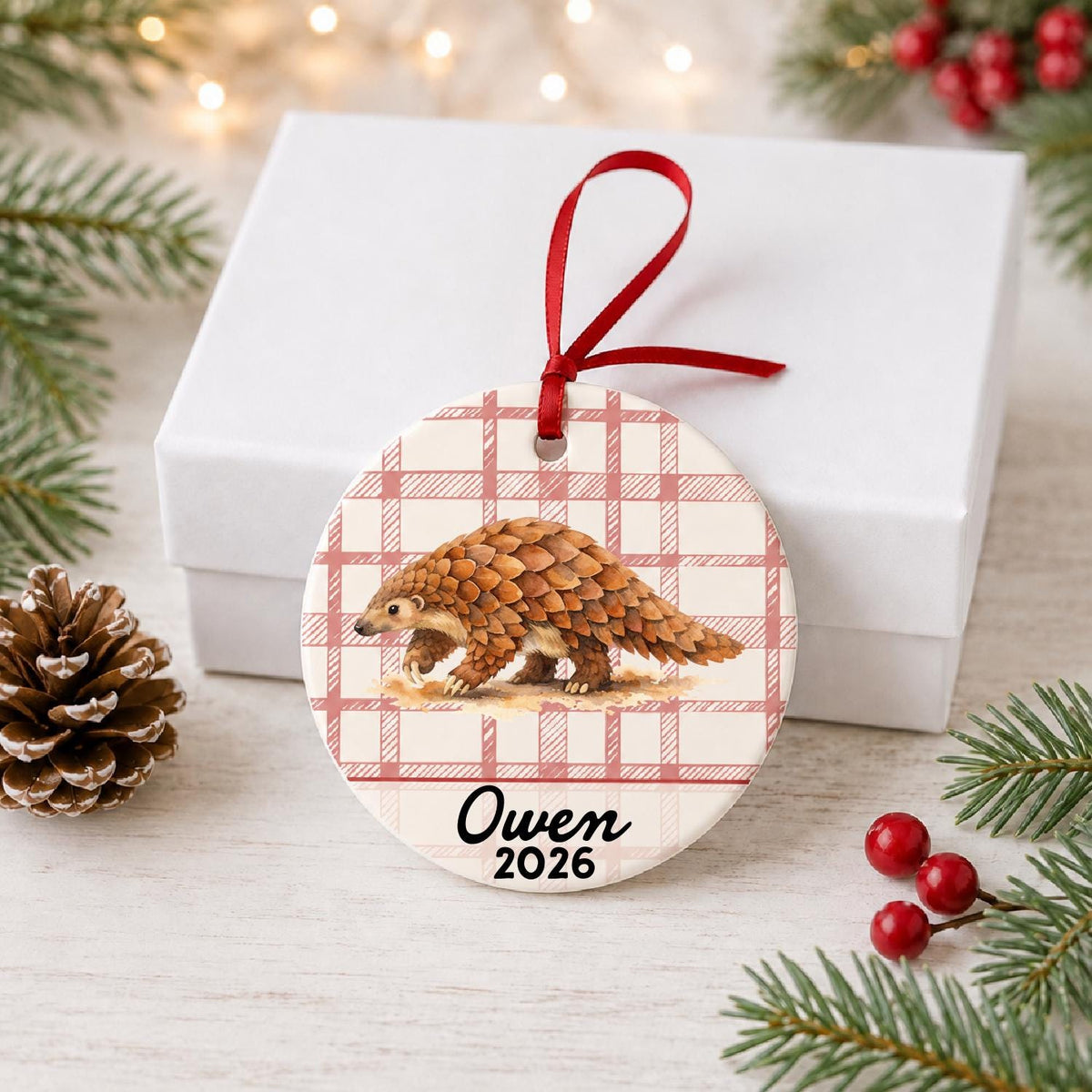 Personalized pangolin ceramic ornament with name and year on red plaid in front of a white gift box with pinecones and red berries
