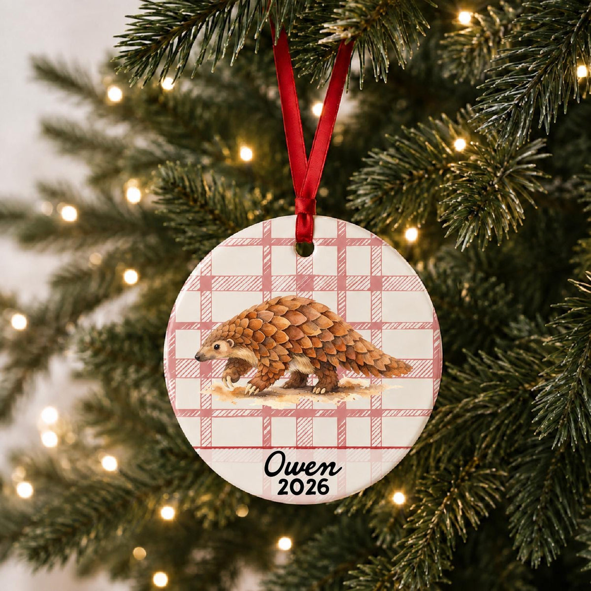 Personalized pangolin ceramic ornament with name and year on red plaid hanging on a lit Christmas tree