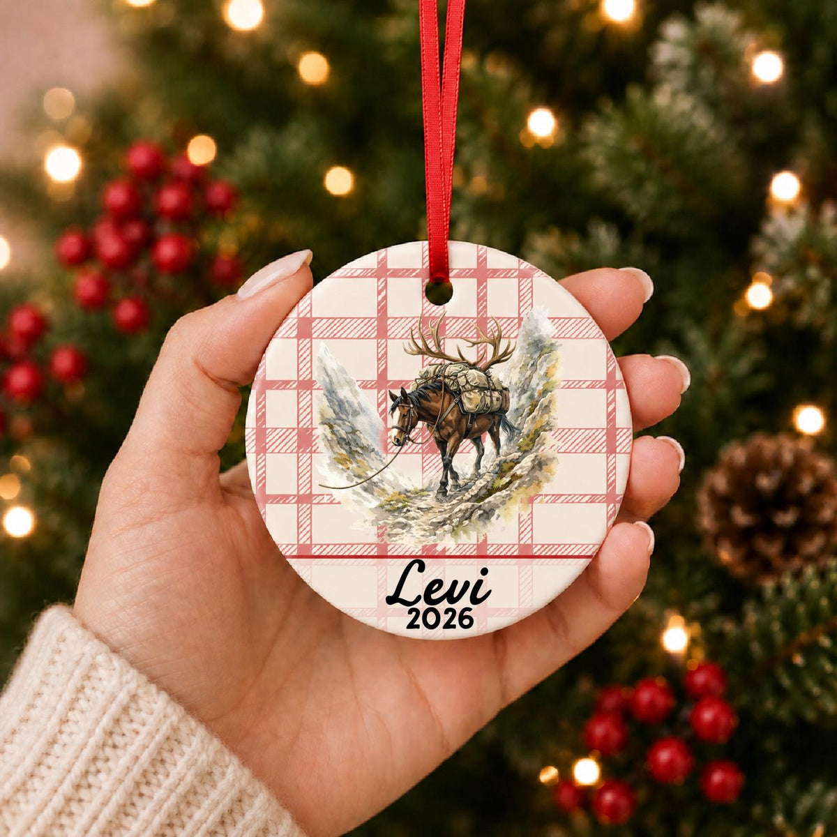 Hand holding the personalized pack horse ceramic ornament with name and year on red plaid in front of a Christmas tree