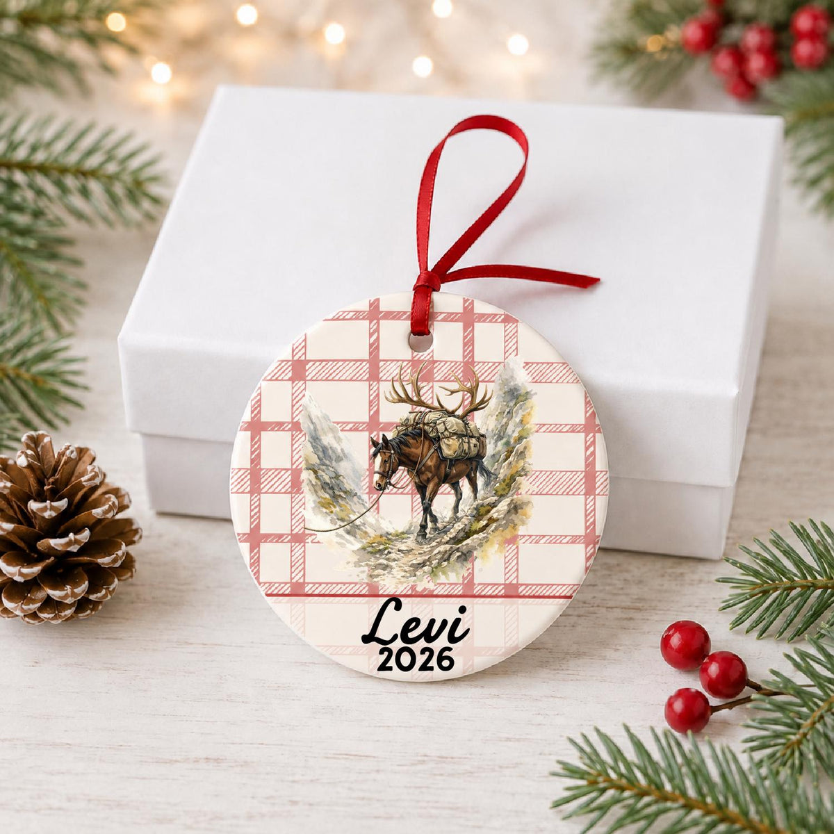 Personalized pack horse ceramic ornament with name and year on red plaid in front of a white gift box with pinecones and red berries