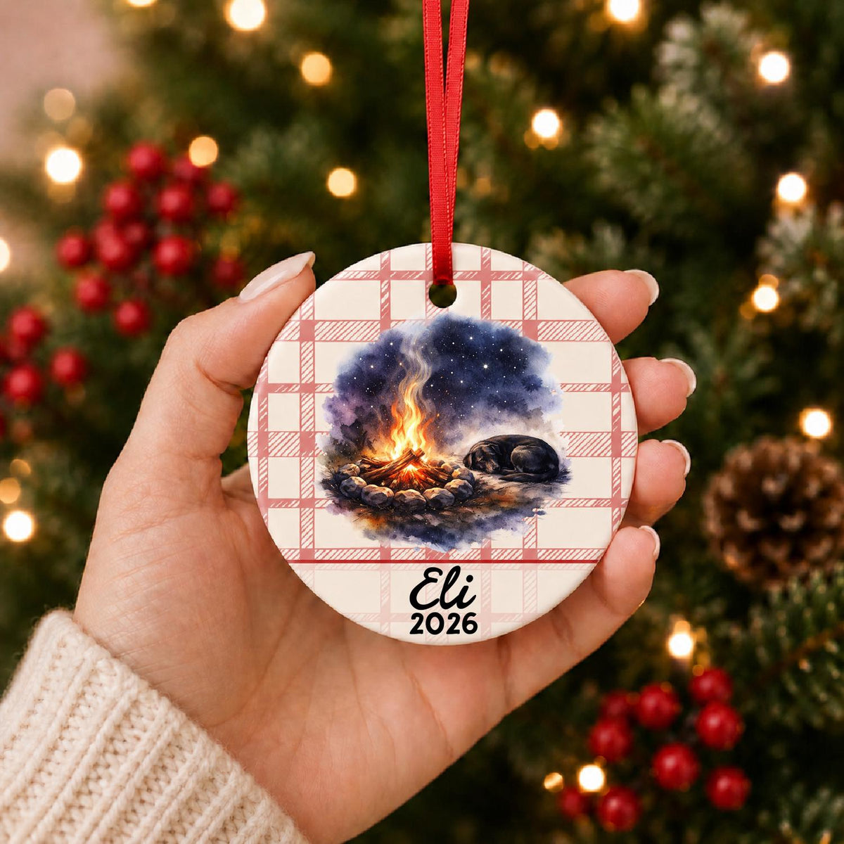 Hand holding the personalized campfire and dog ceramic ornament with name and year on red plaid in front of a Christmas tree
