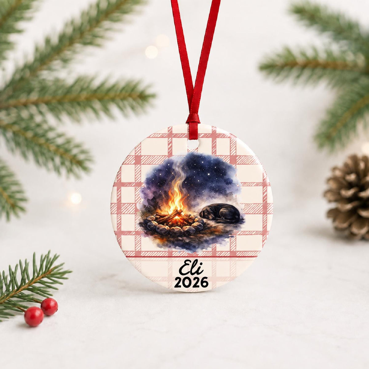 Personalized campfire and dog ceramic ornament with name and year on red plaid with red ribbon on a white surface with pine branches and pinecones