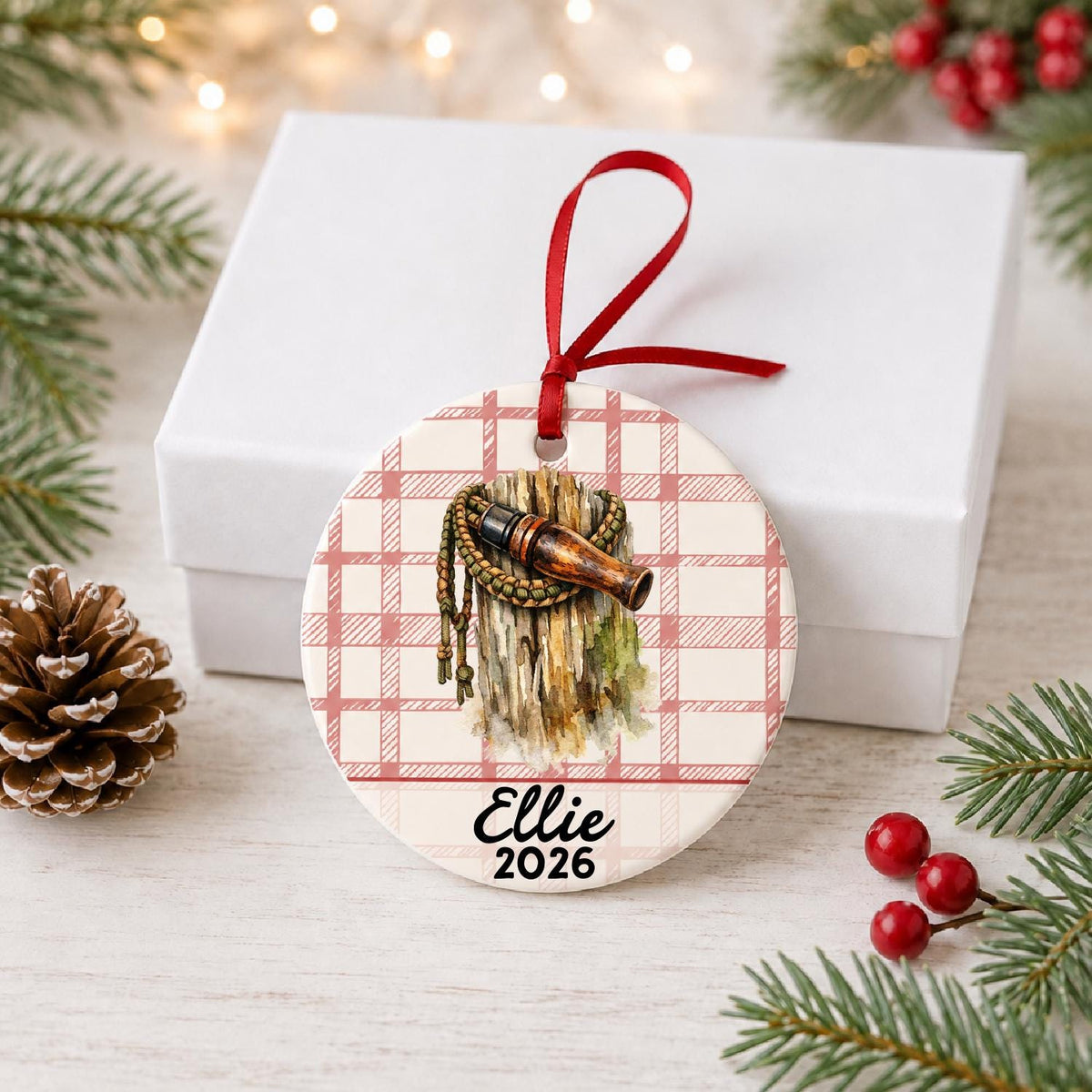 Personalized duck call ceramic ornament with name and year on red plaid in front of a white gift box with pinecones and red berries