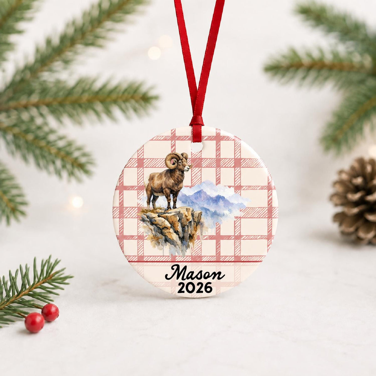 Personalized bighorn sheep ceramic ornament with name and year on red plaid with red ribbon on a white surface with pine branches and pinecones