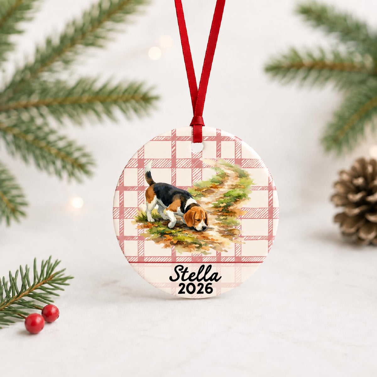 Personalized beagle ceramic ornament with name and year on red plaid with red ribbon on a white surface with pine branches and pinecones