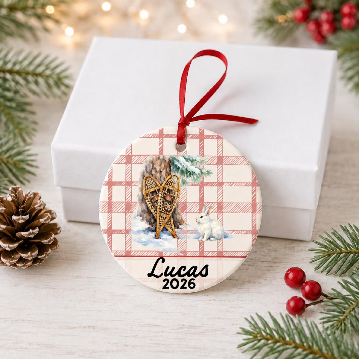 Personalized snowshoes and snowshoe hare ceramic ornament with name and year on red plaid in front of a white gift box with pinecones and red berries