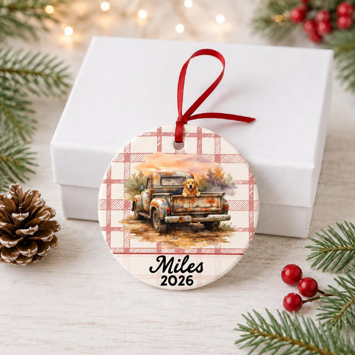 Personalized vintage truck and dog ceramic ornament with name and year on red plaid in front of a white gift box with pinecones and red berries