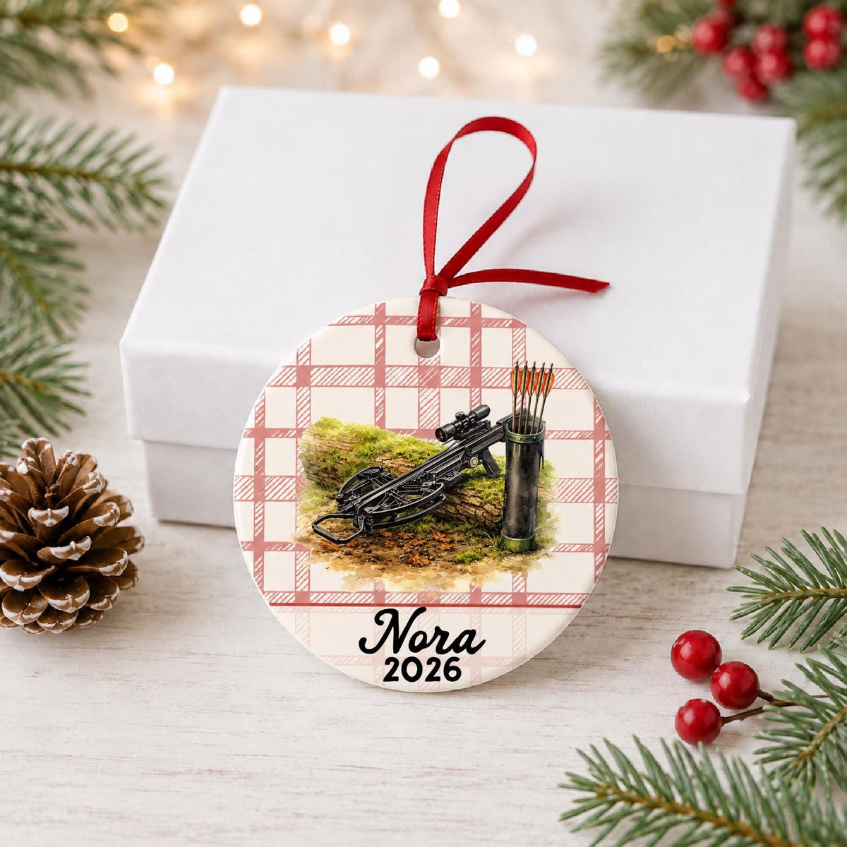 Personalized crossbow ceramic ornament with name and year on red plaid in front of a white gift box with pinecones and red berries