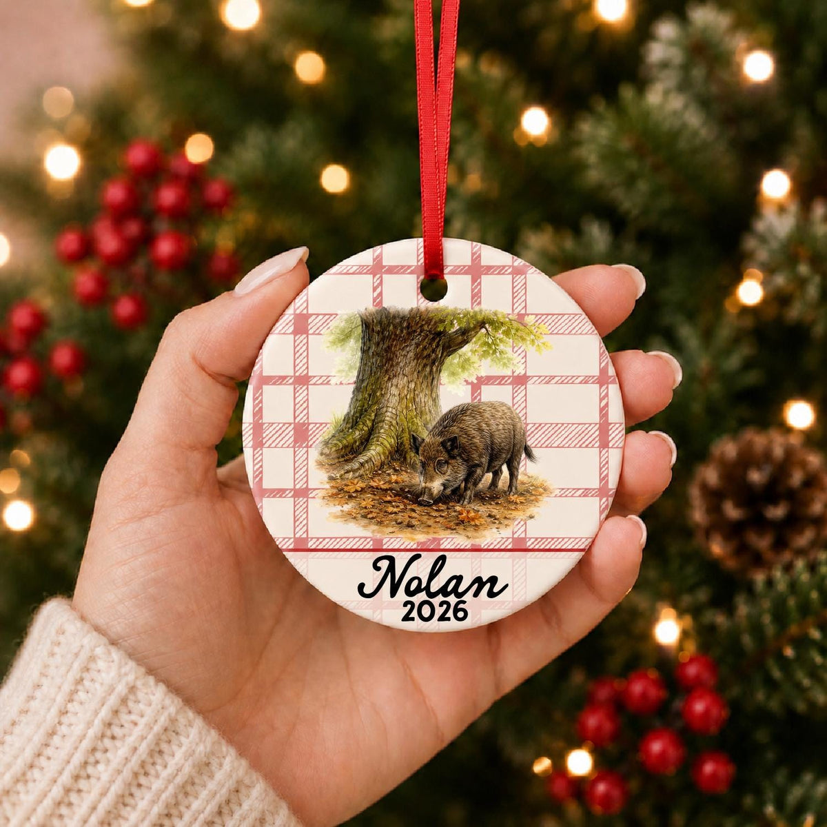 Hand holding the personalized wild boar ceramic ornament with name and year on red plaid in front of a Christmas tree